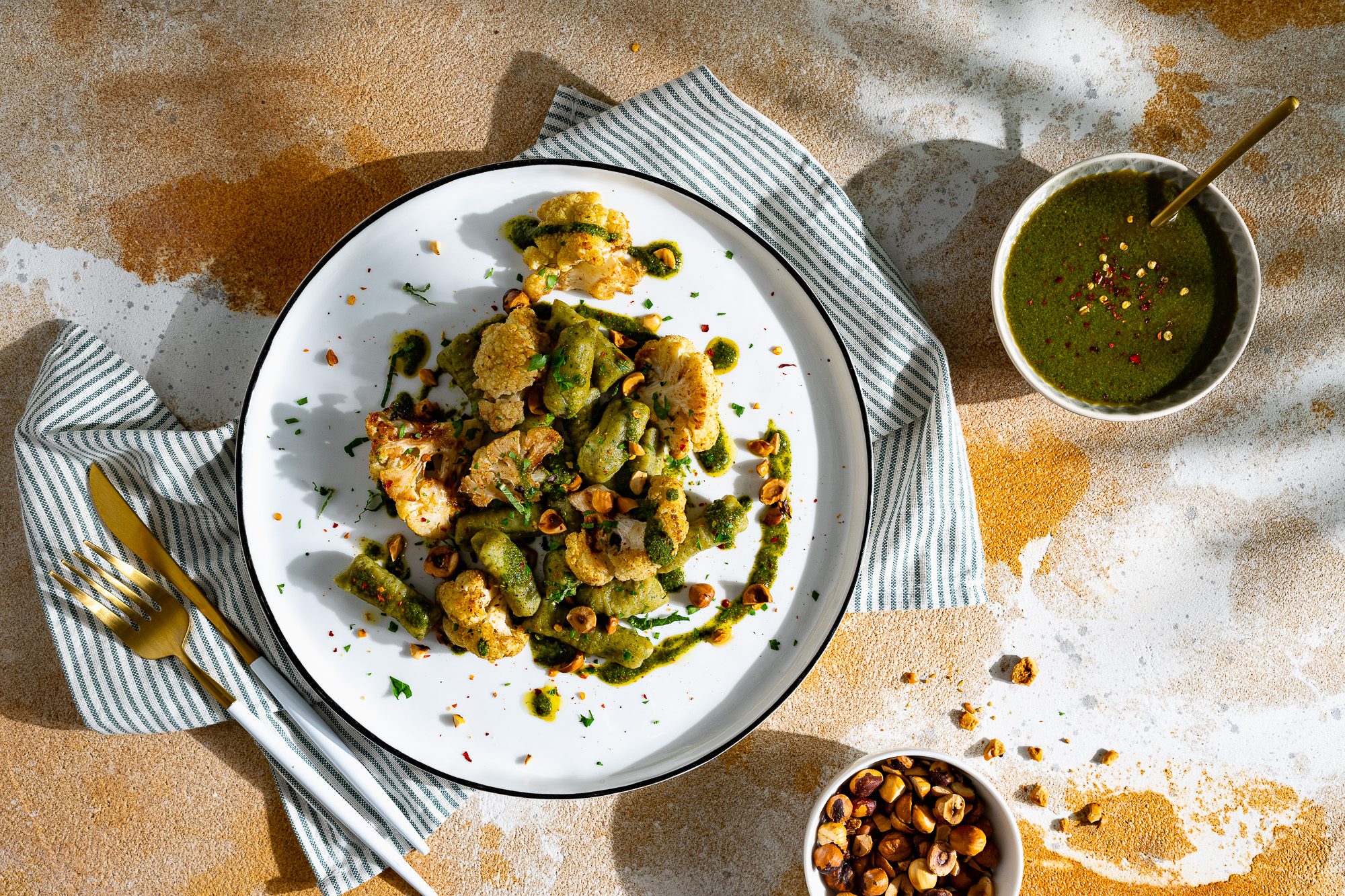 Linsen-Gnocchi mit geröstetem Blumenkohl und Zhug auf einem Teller serviert. Daneben steht ein Schälchen mit Zhug und gerösteten Haselnüssen.