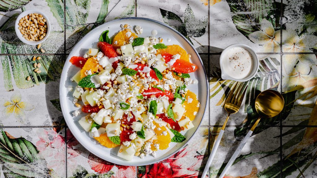 Honigmelonensalat mit Tahini-Joghurt-Dressing auf einem Teller serviert. Daneben steht eine kleine Schale mit gerösteten Pinienkernen und ein Schälchen mit Dressing.