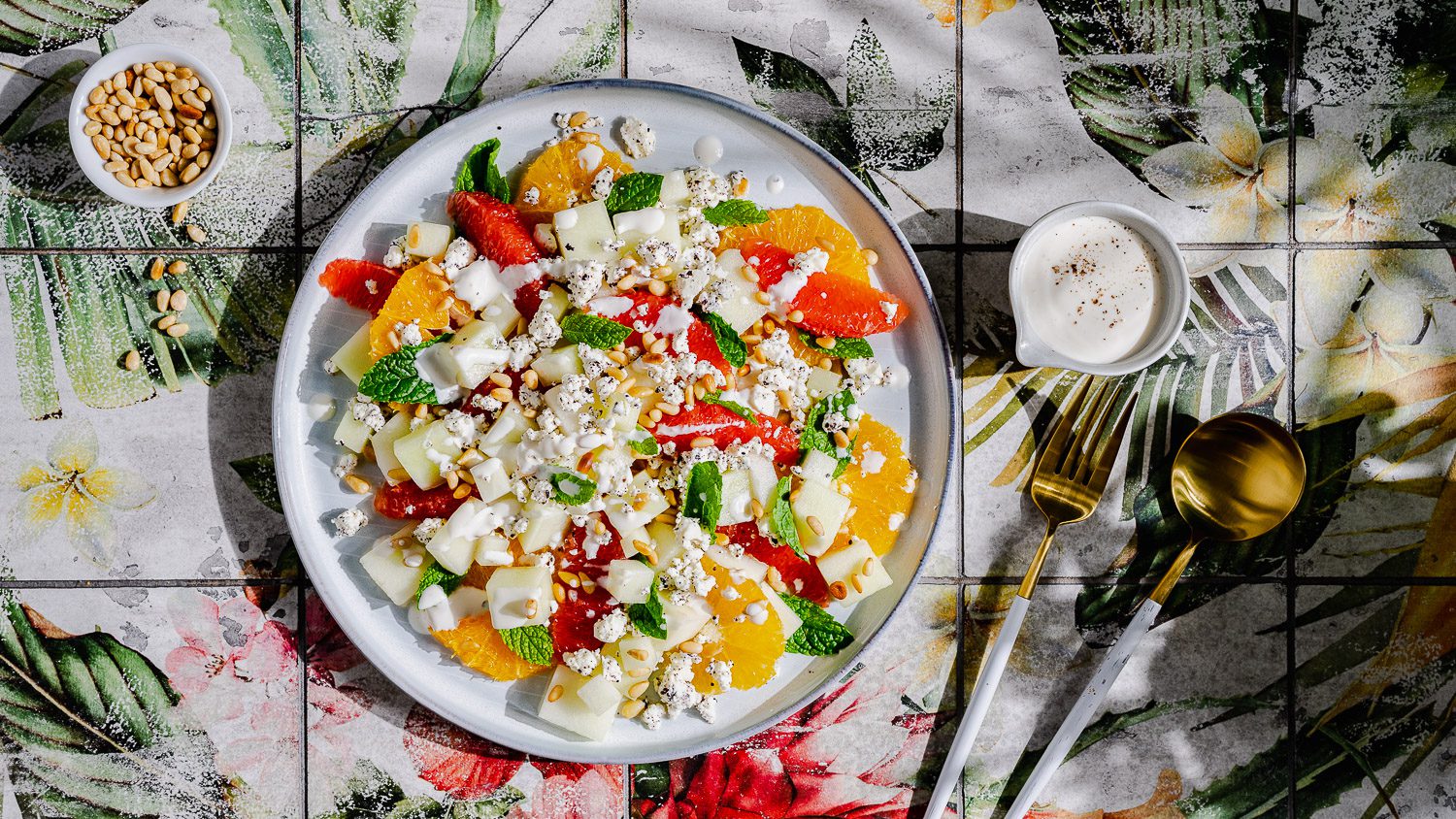 Honigmelonensalat mit Tahini-Joghurt-Dressing auf einem Teller serviert. Daneben steht eine kleine Schale mit gerösteten Pinienkernen und ein Schälchen mit Dressing.