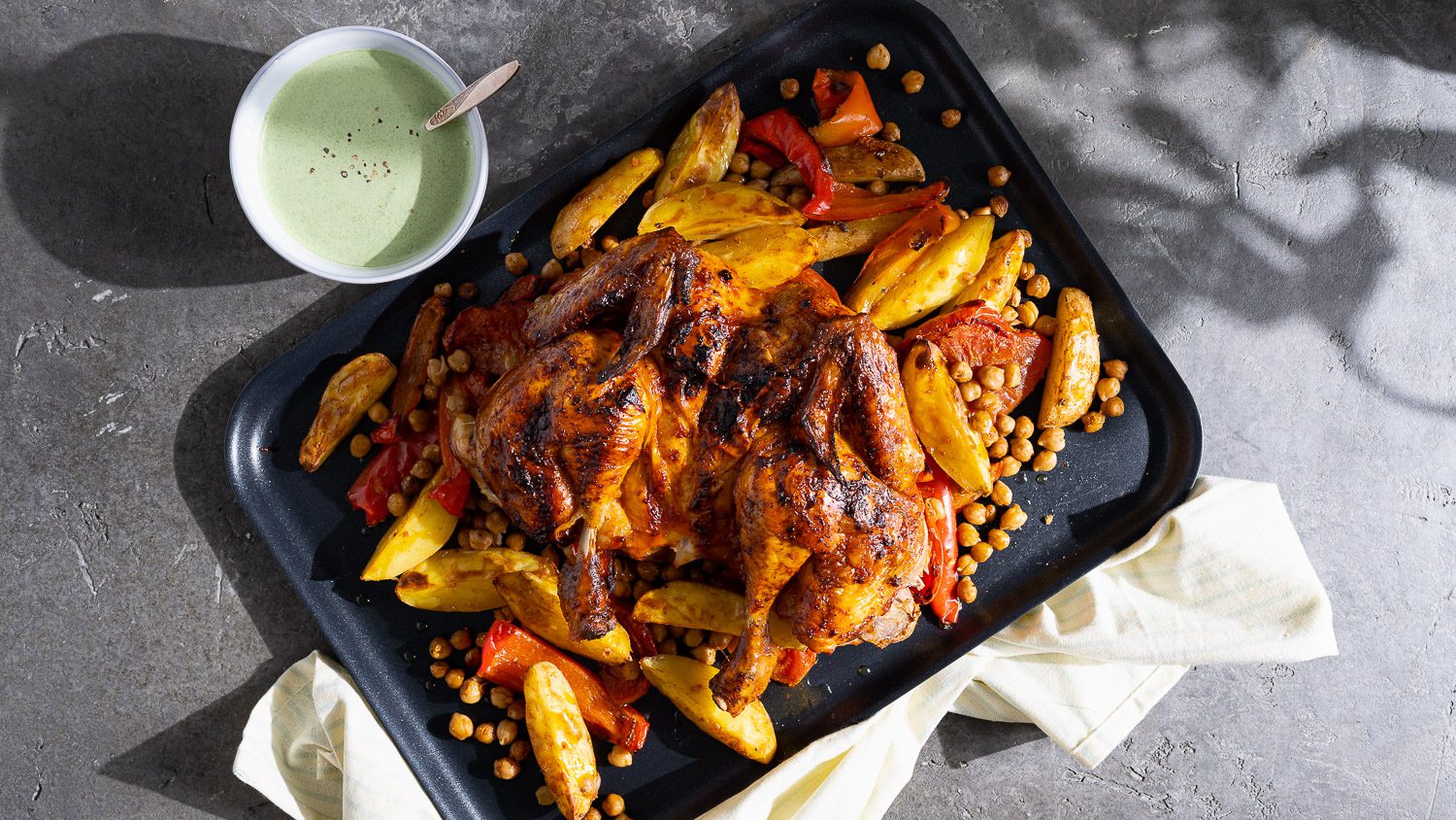 Harissa-Hähnchen mit Ofengemüse, serviert auf einem Backblech. Daneben ein Schälchen Minzsauce.