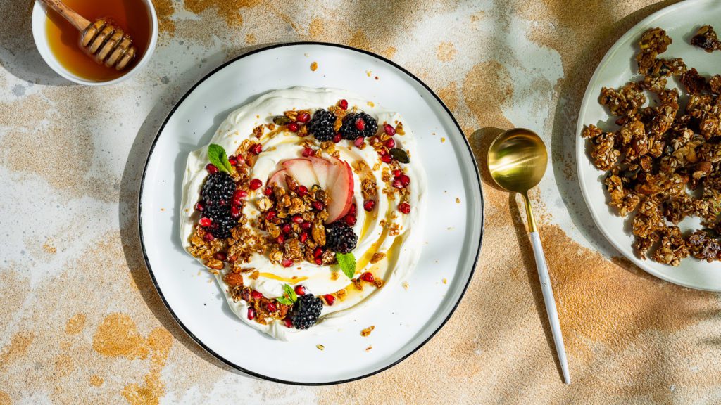 Labneh mit Granola und frischen Beeren auf einem Teller serviert. Daneben stehen noch Honig und Granola.