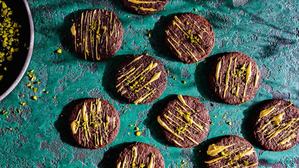 Schoko-Cookies mit Mokka und Pistazien verziert liegen auf einer Tischplatte.