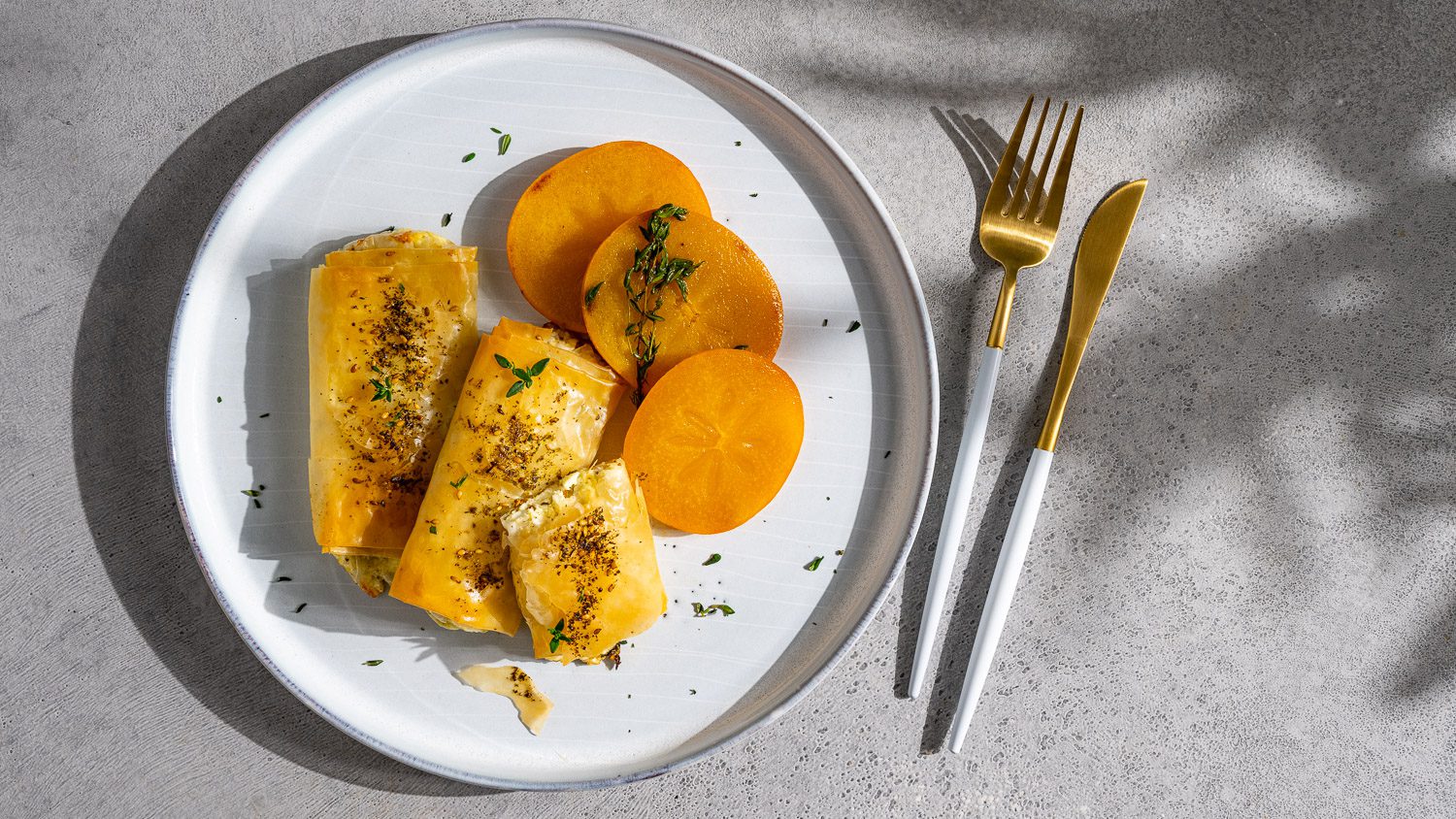 Feta-Päckchen mit Thymian serviert an karamellisierten Kaki auf grauem Untergrund.