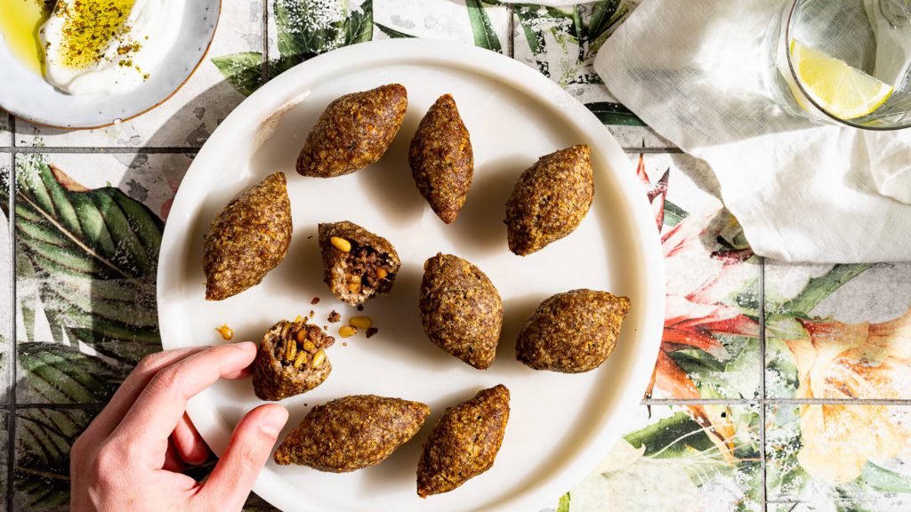 Klassische Kibbeh Kroketten mit Hackfleischfüllung liegen auf einem runden Marmorteller. Eine Kibbeh ist halbiert und wird von einer Hand gegriffen.