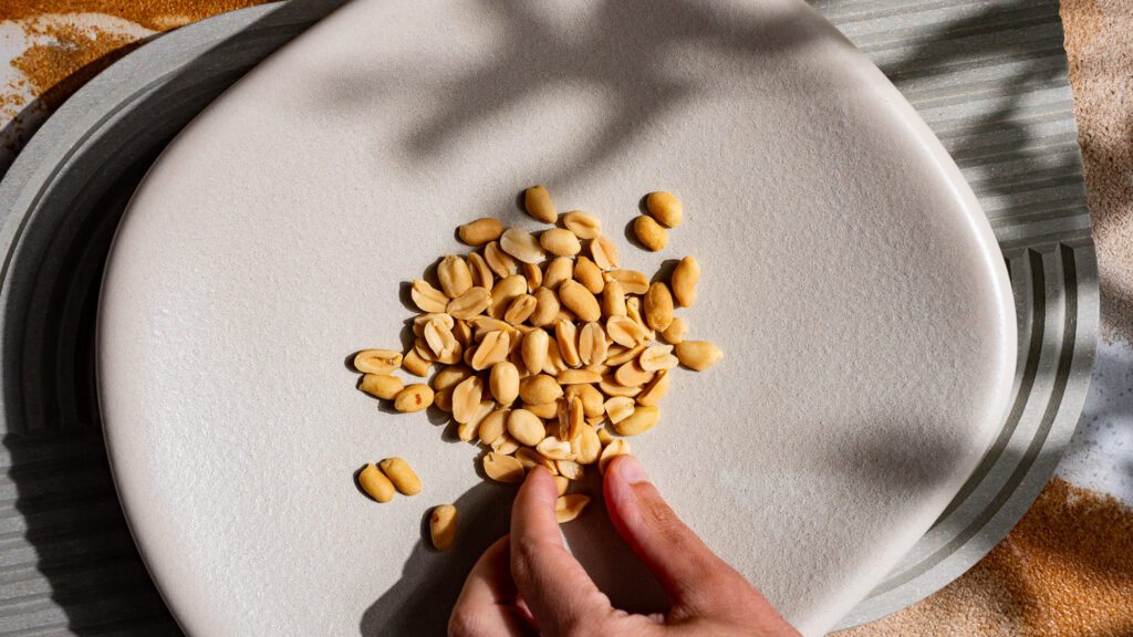 Hülsenfrüchte: Erdnüsse auf einem hellen Teller. Eine Hand greift nach einer Erdnuss.
