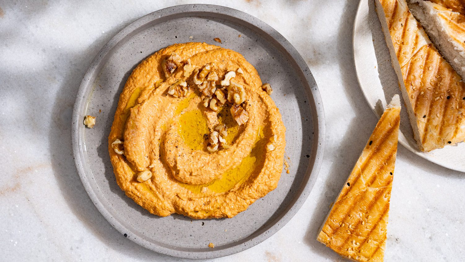 Muhammara, ein levantinischer Paprika-Walnuss-Dip, verstrichen auf einem grauen Teller. Dazu wird Brot serviert.