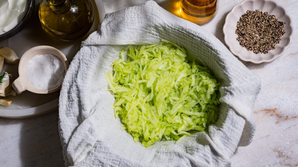 Se pone un pepino rallado en un paño para colar. A su alrededor hay otros ingredientes para el tzatziki.