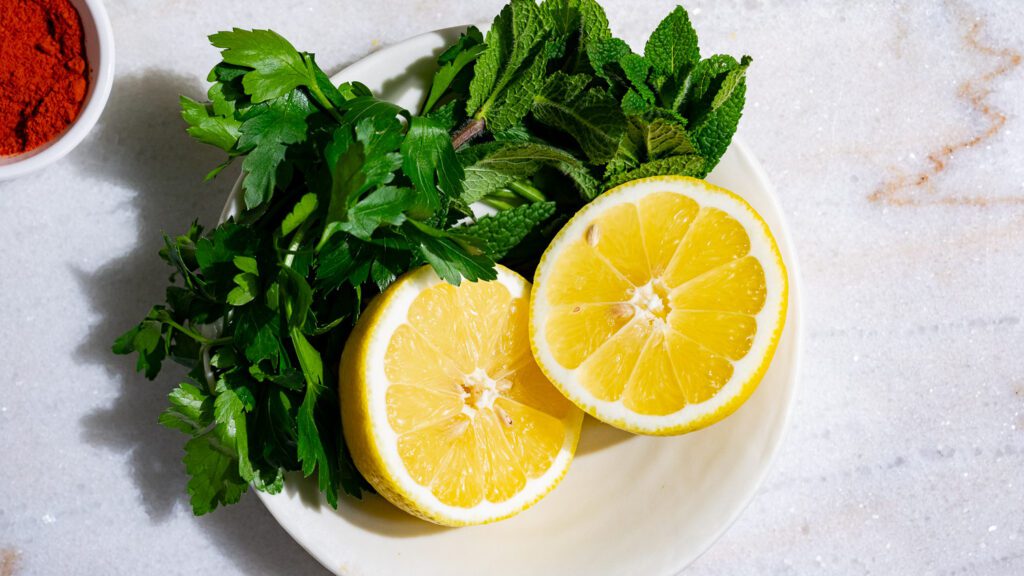 Lemon halves, flat-leaf parsley and mint in a small bowl on a light marble background.