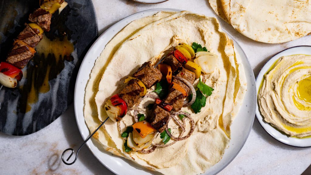 Mesa puesta con shish kebab, brochetas sobre pan plano y hummus.