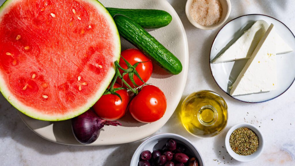 Todos los ingredientes de la ensalada griega con sandía están dispuestos sobre una mesa.