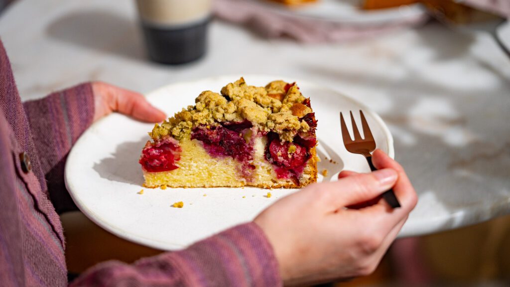 Pflaumenkuchen mit Streuseln auf einem Teller, der gerade gegriffen wird. In der anderen Hand eine Gabel.