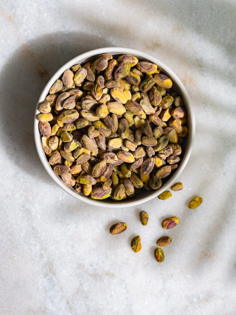 Pistachos en un cuenco sobre una mesa de mármol blanco a la sombra de una planta.