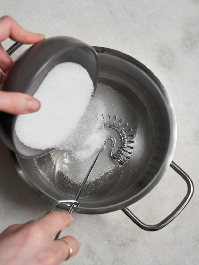 Zucker wird zu Wasser und Schneebesen in den Topf gegeben für den Sirup des Pistazien-Baklavas.