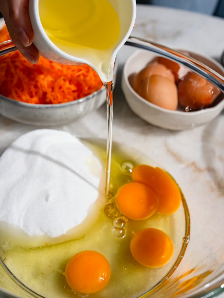 Neutrales Öl fliest in eine Schale mit Zucker und aufgeschlagenen Eiern für einen saftigen Karottenkuchen.