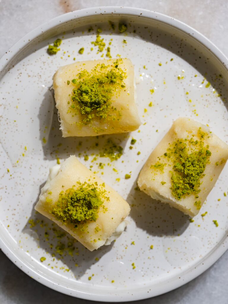Tres trozos de halawet el jibn adornados con pistachos y jarabe de azúcar servidos en un plato.